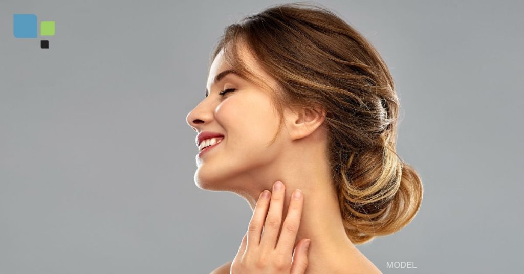 A white woman model with brown hair pulled back into a low bun. Her neck and face is showing in this picture and is turned to the side, with her hand partially resting against her neck.