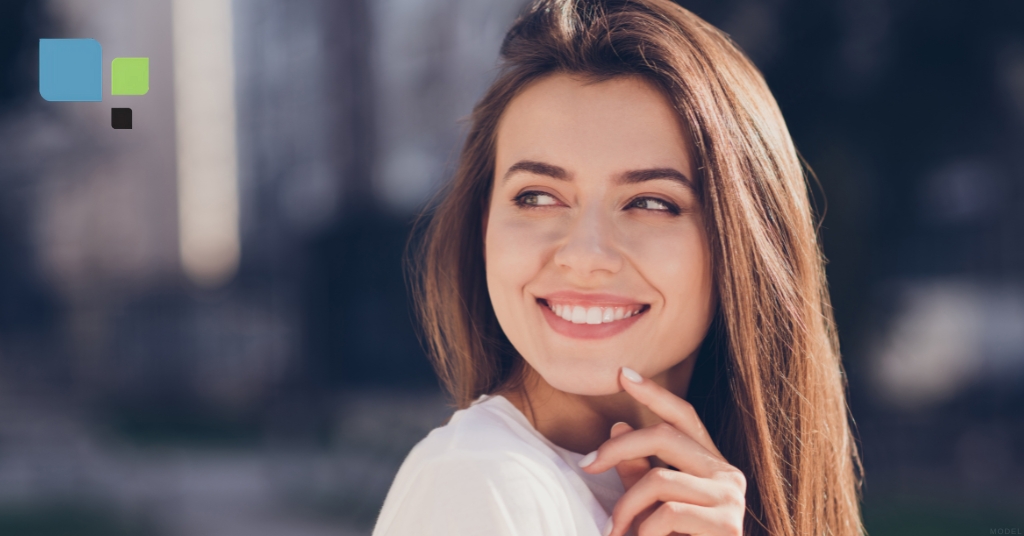 A young woman smiling in the park (model) Primera Plastic Surgery Logo